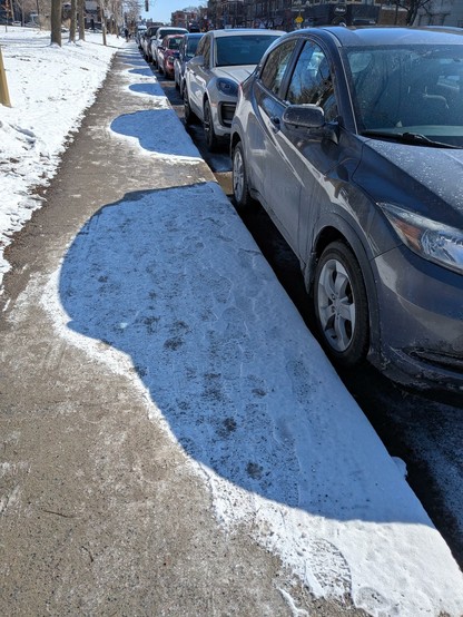 Parked cars are casting shadows on the sidewalk where it had just snowed. The snow is not melted in the shadows, but is in the sun.