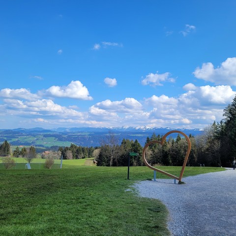 Wanderweg mit weitem Blick in Berge. Himmel mit vielen Wolken. Am Wegrand im Gras ein Herzrahmen im Gras.