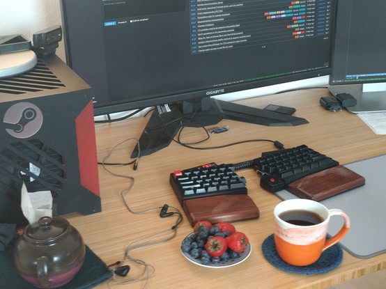 A desk top with a split keyboard, a small plate with blueberries and strawberries, and a cup with black tea. 

In the background there is a computer case with the steam logo and the bottom of a monitor which shows a list of tickets on the GoActivityPub bug tracker.