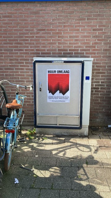 Leeg reclame frame op elektriciteitskastje met poster: Bovenaan staat in grote zwarte hoofdletters “HUUR OMLAAG”, daaronder, op de achtergrond een graphic van twee omgedraaide, rode rijtjeshuizen, de punten van de daken wijzen omlaag. Op de voorgrond staat een tekst in witte letters: Landelijke demonstratie in Utrecht, Zaterdag 10 mei, 14.00, Domplein, woonprotestutrecht.nl 
Blauwe fiets met bruin zadel ernaast geparkeerd. Bakstenen muur.