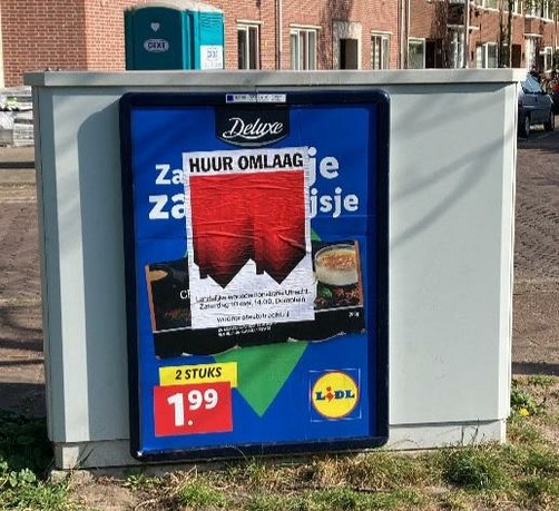 poster over reclemae bord op kastje: Bovenaan staat in grote zwarte hoofdletters “HUUR OMLAAG”, daaronder, op de achtergrond een graphic van twee omgedraaide, rode rijtjeshuizen, de punten van de daken wijzen omlaag. Op de voorgrond staat een tekst in witte letters: Landelijke demonstratie in Utrecht, Zaterdag 10 mei, 14.00, Domplein, woonprotestutrecht.nl
Op de achtergrond woningen en een dixie