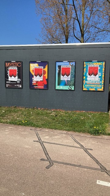 posters geplakt op 4 reclame borden op huisje met donker geverfde bakstenen, Bovenaan staat in grote zwarte hoofdletters “HUUR OMLAAG”, daaronder, op de achtergrond een graphic van twee omgedraaide, rode rijtjeshuizen, de punten van de daken wijzen omlaag. Op de voorgrond staat een tekst in witte letters: Landelijke demonstratie in Utrecht, Zaterdag 10 mei, 14.00, Domplein, woonprotestutrecht.nl
Boom achter en blauwe lucht, voor gras en fietspad