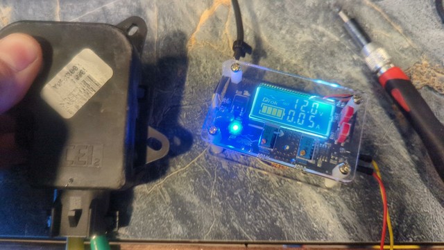 The old blend door actuator. It's a small black plastic housing, with a faded white sticker attached. The output shaft isn't visible, and is being held down (prevented from turning) by a finger and thumb. To the right, a small adjustable power supply shows 12Vdc and .05A as the motor tries to turn against the resistance.