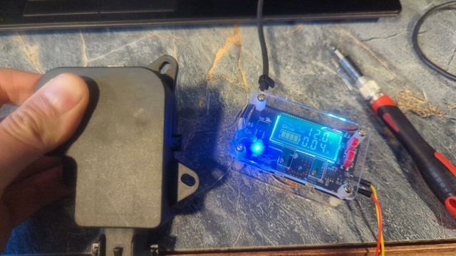 The new blend door actuator. It's a small black plastic housing, much cleaner than the old actuator. The output shaft isn't visible, and is being held down (prevented from turning) by a finger and thumb. To the right, a small adjustable power supply shows 12Vdc and .04A as the motor tries to turn against the resistance.