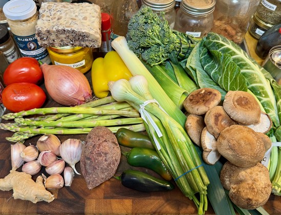 A cutting board which are seen a jar of Bubbies horseradish, a block of tempeh, a jar of tumeric paste, two Roma tomatoes a large shallot, about 8 spears of asparagus, 9 cloves of red garlic, a section of ginger root, a purple potato, 3 jalapeños, 3 cambray onions, a leek, 1/2 large yellow bell pepper, 2 leek tops (the green leafy part), one bunch broccolini, 1 bunch collars and a pile of shiitake mushroom caps.