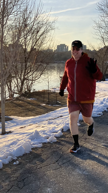 À man in a red jacket and orange shorts and black ball cap waves at the camera as he runs along the trail in a snowy-melty park.