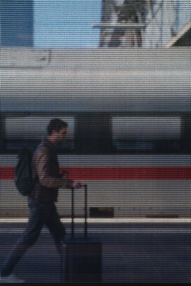 Traveler walking on a platform, photographed through the sunscreen of a train car. Behind the traveler, another train is visible.