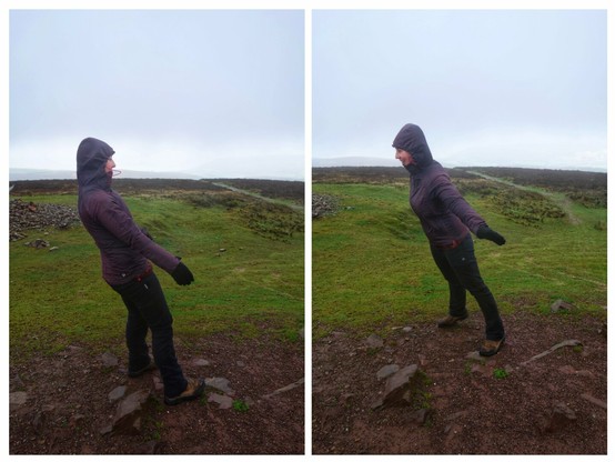 Two side-by-side images show a woman standing on a rocky, muddy ground in a windy outdoor setting. In both images, the woman is wearing a dark purple hooded jacket, dark trousers, gloves, and brown hiking boots. The hood of her jacket is up, covering her head.
In the left image, the woman is leaning significantly to her left, with her arms extended outwards for balance. Her body is angled, and she appears to be bracing against a strong wind coming from her right. Her gaze is directed slightly downwards and to her left.
In the right image, taken from a slightly different angle, the woman is leaning to her right, again with her arms extended for balance. She is smiling and looking towards her left, suggesting she is interacting with someone or something off-camera.
The background in both images is a landscape of rolling green hills covered with patches of dark vegetation. A faint, hazy horizon is visible under a grey, overcast sky, indicating potentially damp or misty weather conditions.