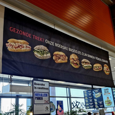 Een reclamebanner in een winkel met de tekst "Gezonde trek? Onze Boerske broodjes zijn binnen verkrijgbaar!" en afbeeldingen van verschillende vettig ogende belegde broodjes. Onder de banner hangt een bord met de tekst "Boerske is cashless - hier alleen pinnen." Rechts naast het bord is een menukaart en een aanbieding voor een "combi deal" zichtbaar.