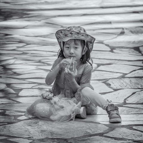 Young girl sitting on sidewalk pauses from playing with her doll to take a sip of her drink.