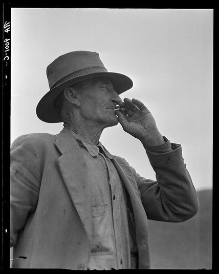 The image is a black and white photograph depicting an elderly man standing outdoors. He wears a wide-brimmed hat, possibly made of straw or similar material, with what appears to be decorative perforations around the edge. The man's expression seems contemplative as he looks off into the distance while holding his pipe near his mouth.

He is dressed in casual work attire consisting of a worn suit jacket and trousers that suggest labor-intensive activity; there are visible stains on the cuffs indicating manual or outdoor work. His shirt appears to be buttoned up, revealing pockets where tools or personal items could potentially have been stored during his day's activities.

The backdrop is nondescript but seems open-air with soft lighting conditions suggesting an overcast sky or a diffuse light source at either dawn or dusk. The photo has no visible contextual elements that provide further information about the man’s identity, location beyond California (as indicated in the caption), or any specific event he might be involved in.

The photograph is identified as part of Dorothea Lange's collection and carries her name on it; however, she was not a labor contractor but rather an American photographer known for documenting social issues. The image captures a moment reflective of working-class life during that era with a focus on the man’s perspective or viewpoint—implied through his thoughtful posture and gaze.

The captio [...]