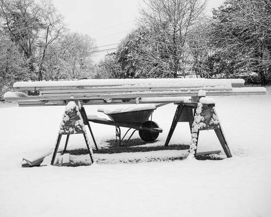 Sawhorses holding lumber with a wheelbarrow underneath. Monochrome