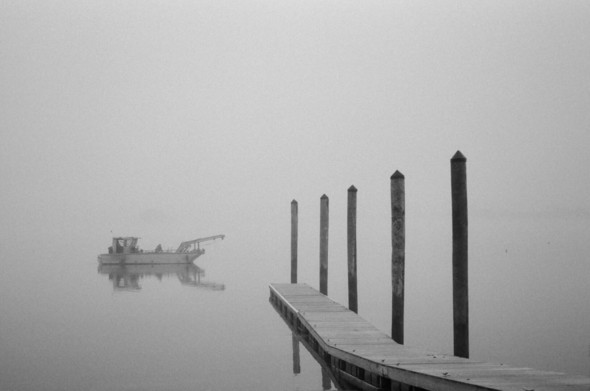 A small utility craft floats off the end of a pier amidst a fog that obscures the border between water and sky.