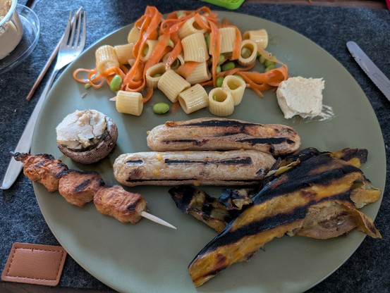 Ein grüner runder Teller, darauf auf der unteren Hälfte links ein Spieß & ein gefüllter Champignons, mittig zwei Bratwürste, rechts gegrillte Auberginenscheiben. Auf der oberen Hälfte Nudelsalat und rechts noch ein Klecks Hummus