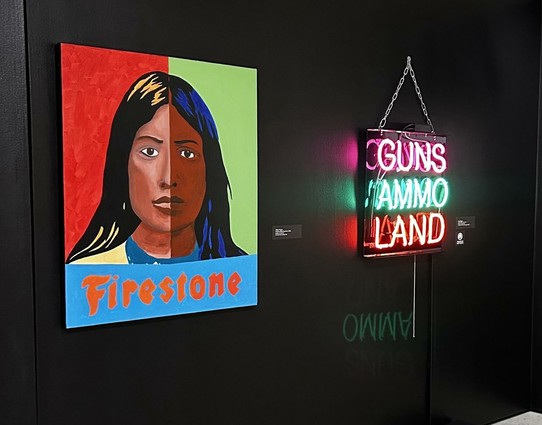 Two works hanging on a black wall. Painting of a face vertically split, and the word Firestone.
Neon words : GUNS AMMO LAND.