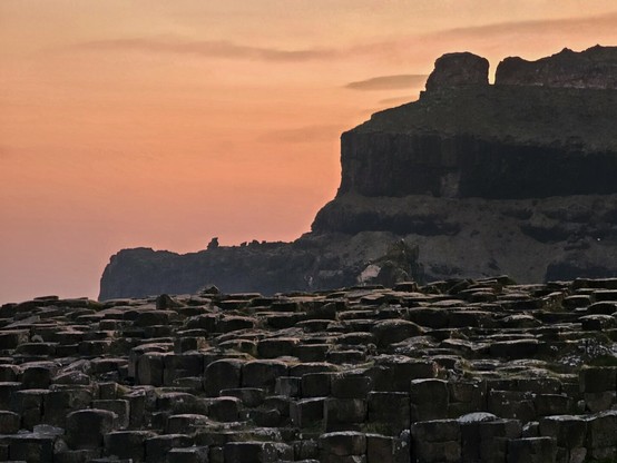 A wide shot shows the hexagonal basalt columns of the Giant's Causeway leading out to the sea, with a dramatic cliff face in the background under a soft, orange-tinged sky.