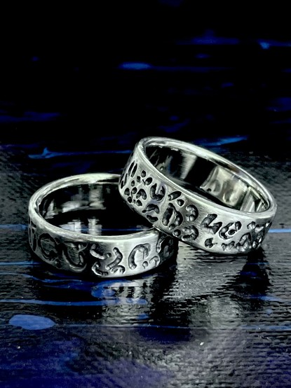 Two handmade silver rings are shown, one featuring a smooth surface and the other with a textured, patterned design. The rings rest on a dark, reflective surface that enhances their shine.
