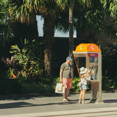 The image shows a man and a child standing near a public phone booth. The man is holding shopping bags and the child is interacting with the phone. The booth has a Telstra logo on top. They are surrounded by tropical plants and trees, suggesting a warm climate setting.