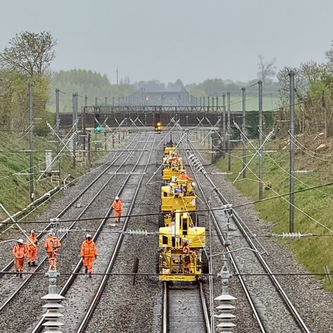 Photo of men working on the tracks of a railway. A group of men in high visibility overalls are walking towards the foreground on the second of four lines. On the third line is a line of yellow maintenance cars. Electric cables cross the photo and ceramic insulation can be seen at the very front if the image. A footbridge crossing the lines is in the background.