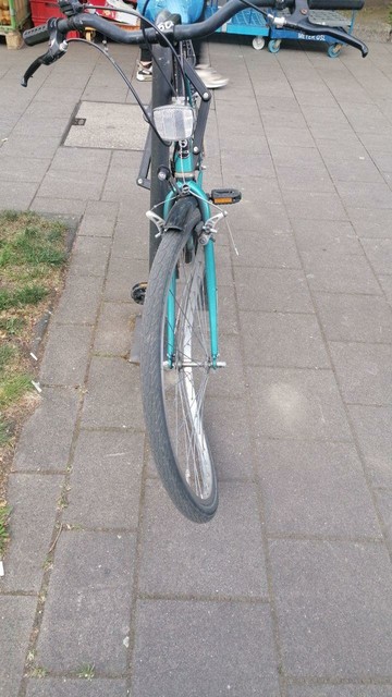 Ein angeschlossenes Fahrrad (grün /blau) von vorne mit schwarzem Lenker. Das Vorderrad ist verbogen. Im Hintergrund Paletten. Untergrund: große Pflastersteine