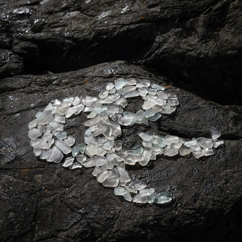 An ampersand made by arranging white sea glass on a dark grey rock. The glass glistens after a bit of a splash by a nearby wave, in the sunlight.