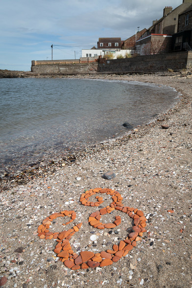 An ampersand made by arranging worn terracotta tile remnants on the beach of a sheltered cove.