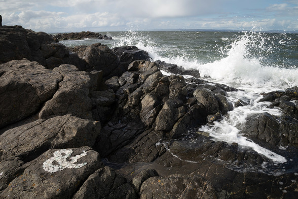 An ampersand made by arranging white sea glass on a dark grey rock. The waves crash on the rocks, in the background.