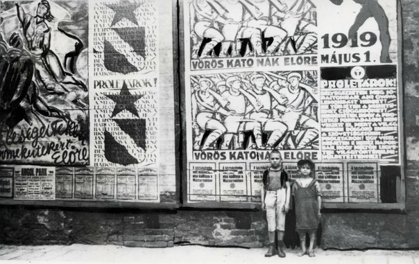 A black and white photo of two children standing up against a large wall, possibly the side of a building, that features giant posters with depictions of soldiers, as well as a lot of writing. The children are dressed in poor clothing, suggesting they live in poverty. One is a boy, older and taller than the girl standing beside him. Both are looking directly at the camera and are standing one-third to the right of the frame, illustrating perfectly 'the rule of thirds'.