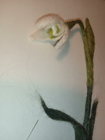 On a white background lies a snowdrop made of nettle using needle felting. This will be a bookmark in the form of a plant. Thank you for your attention!