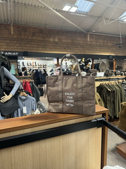 A brown tote bag with the text "ENJOY THE GAME" displayed prominently. The bag is positioned on a wooden display in a retail store, with clothing racks and merchandise visible in the background.