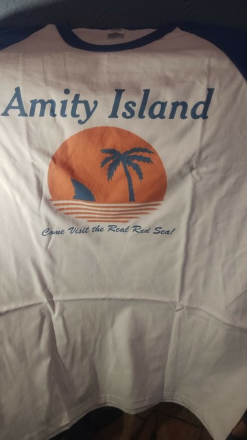 Two tone t-shirt with white body, blue collar and sleeves, stylised sun with palm tree and shark gin silhouette, with "Amity Island" above the image and "come visit the real red sea" below