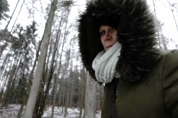 The image shows a person in a winter setting, wearing a thick, fur-lined hooded coat and a scarf. The background consists of tall trees in a snowy forest.