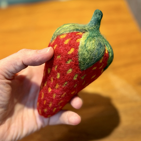 Susan’s hand holds a large red needle felted strawberry with green leaves and yellow/yellow green seeds. Background is a wooden desk top.