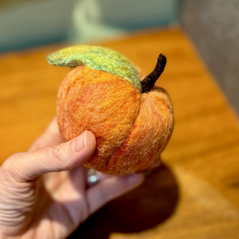 Susan’s hand holds a needle felted peach with a green leaf and brown stem. Background is a wooden desk top.