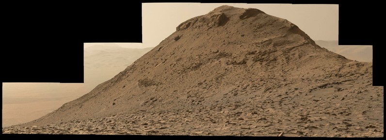 An ochre toned mosaic of 18 images stitched together against a black background, featuring a Martian butte. The wall of the rim of Gale Crater is seen in the background at a distance of 10 kilometers. The images were captured by the Mars rover Curiosity's Mastcam science camera.