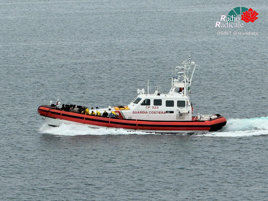 Die GP 323 der Guardia Costiera in gemäßigter Fahrt. Auf dem Vordeck gedrängt Menschen, teilweise in Rettungsfolien gehüllt.

Gekennzeichnet mit Radio Radicale Logo, OSINT @scandura