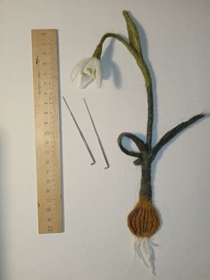 On a white background near the line lies a snowdrop, as if it were being pulled out of the ground. This is a bookmark with a picture of plants, made of nettle with needle felting. Next to the snowdrop, leave two special toys for felting. Thank you for your attention!
