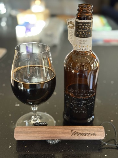 A snifter glass of dark beer next to a tall brown bottle with a white neck label with gold lettering. The bottle is embossed with the Goose Island logo. A wooden opener is placed in front.