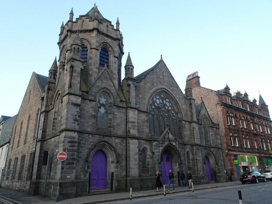 Una iglesia en la esquina de una calle, en la parte antigua de una ciudad. Muros de piedra gris, puertas pintadas de morado. Algunas personas caminando por la acera.