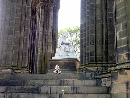 Una gran escultura de un hombre sentado, de mármol blanco, sobre un pedestal, entre enormes columnas (arcos) de piedra negruzca. En el escalón superior de la escalera que sube hasta la parte del monumento donde se encuentra la estatua, hay una niña sentada con las piernas cruzadas y la cabeza apoyada en las manos.