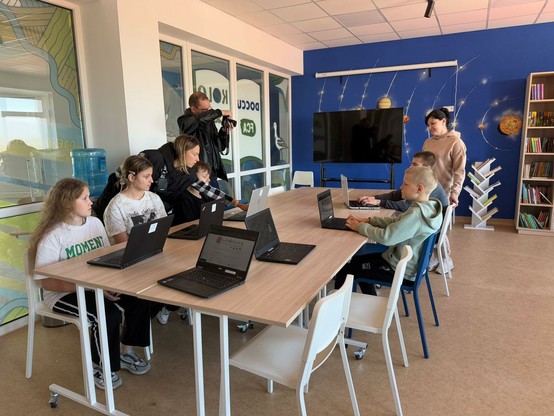 An 4 zusammen gestellten Tischen sitzen 5 Schüler und Schülerinnen auf weißen Stühlen Neben dem Tisch steht ein Mann mit Fotoapparat und eine Frau. Auf dem Tisch stehen 7 aufgeklappte Laptops. Der Raum ist ein Schulzimmer