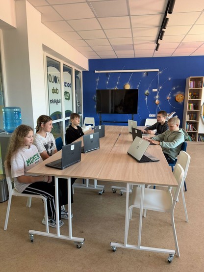 An 4 zusammen gestellten Tischen sitzen 5 Schüler und Schülerinnen auf weißen Stühlen Neben dem Tisch steht ein Mann mit Fotoapparat und eine Frau. Auf dem Tisch stehen 7 aufgeklappte Laptops. Der Raum ist ein Schulzimmer