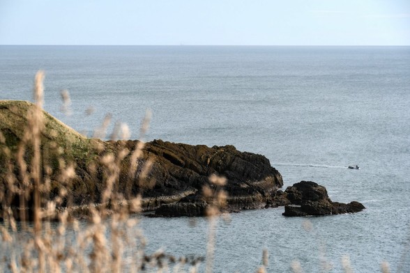 Sicht auf einen felsigen Ausleger der Küste und das offene Meer von weiter oben. Am linken Bildrand sind unscharf Gräser zu erkennen. Außerhalb der Bucht liegt ein kleines Boot. Der Himmel ist blau, das Wasser graublau, die braunen Sandsteinfelsen (denke ich) sind stark verwittert und zur Landseite hin mit Gras bewachsen.