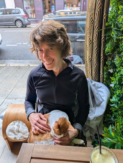 An orange and white cat sits in a woman's lap. The woman is looking at the camera, while the cat is demanding skritches.