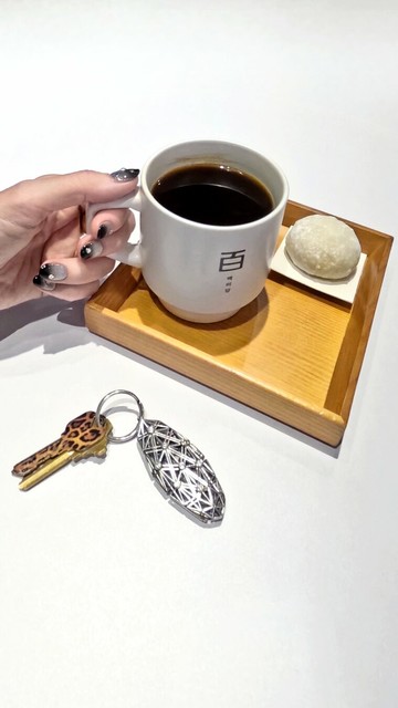 On a white table in a coffee house sits a wooden tray with a cup of hot black coffee and a cold, doughy mochi, beside a silver keychain with bezel-set white sapphires. A hand, its fingernails painted to match the keychain, grasps the coffee mug.