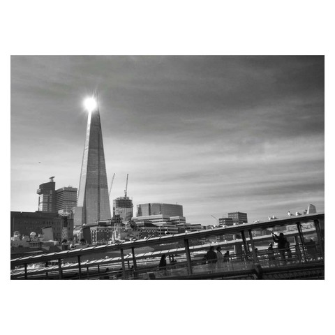 This is a black and white landscape format image of The Shard in London. Taken from across the Thames, in the foreground are the roofs of jetties for boats to land, angled from around a quarter of the way up the frame on the right to the bottom left corner. This angle compliments the tall, thin pyramid shape of The Shard, which sits in the background around a third in from the left, towering above the surrounding buildings. Sunlight is catching and reflecting off the top of The Shard, creating a spotlight effect. Taken with a Sony RX100.