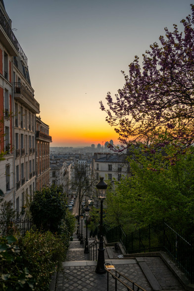 Ein Blick auf Paris von Montmartre aus. Die Sonne geht auf und der Himmel zeigt sich in rotorangenen Tönen. Im Hintergrund sieht man die Stadt mit den hohen Türmen vom Bankenviertel. Links im Bild sind Gebäude zu sehen, rechts Äste eines Baumes. Unten im Foto sieht man eine Treppe, beleuchtet mit den typischen Pariser Laternen, die in die Stadt führt.