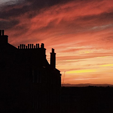 Un edificio antiguo con varias chimeneas de diferentes tamaños, durante una puesta de sol. Los colores del cielo y las nubes son intensos, principalmente rojos y anaranjados, pero también con toques de amarillo, y una parte en gris oscuro en la esquina superior izquierda. Del edificio solo se ve la silueta, casi negra.