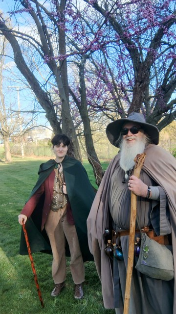 Middle aged man with long grey beard and Gandalf-style outfit with hat and staff wearing sunglasses smiles at the camera.
Just behind is another person, dressed in a Hobbit-Style outfit with dark short hair, and a very stylish cane, looking into the camera.
Behind us is a tree with purplish budding flowers.