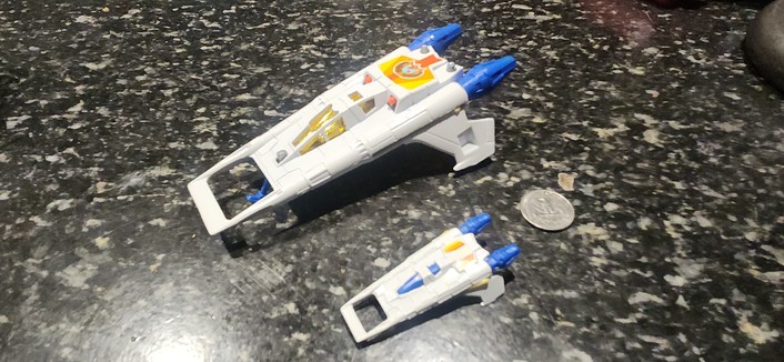 Two white toy spaceships with blue engine nozzles on a speckled black counter top, 25c coin for unhelpful size comparison.